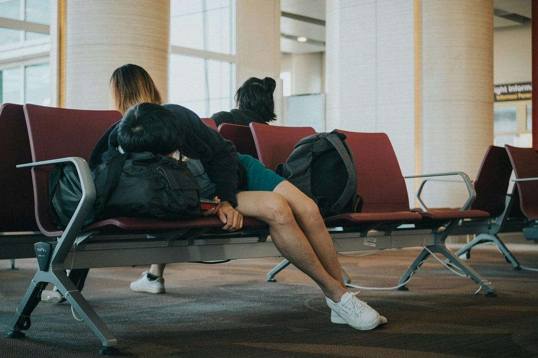 A person sleeps on the chairs at an airport, using their bag as a pillow