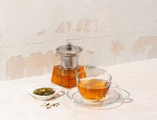 A glass pot of tea, tea leaves in a little dish, and a glass mug and saucer